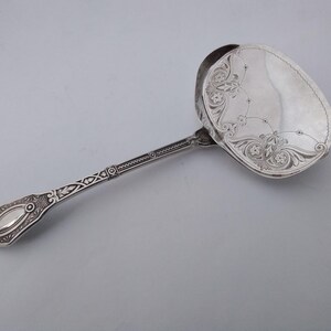 May include: A silver serving spoon with an ornate floral design on the bowl and handle. The spoon has a long, slender handle with detailed patterns. The bowl is oval-shaped and features a decorative floral motif. The spoon is likely made of sterling silver.