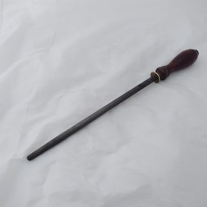May include: A dark grey steel knife sharpening rod with a dark brown wooden handle. The handle has a brass-coloured ring near the steel rod. The rod is designed to sharpen knives.