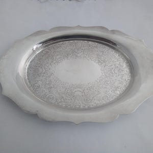 May include: Oval silver serving tray with a decorative, scalloped edge. The tray's surface features an ornate, engraved floral pattern. The tray is made of a shiny, reflective metal.