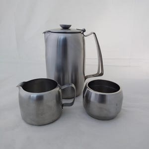 May include: A set of three stainless steel beverage service items consisting of a tall cylindrical coffee or tea pot with a lid and handle, a small creamer jug with a pouring spout and handle, and a matching open sugar bowl container.