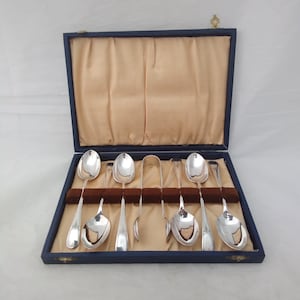 Puede incluir: Un juego de seis cucharas plateadas y unas pinzas para azúcar a juego dispuestas en una caja de presentación rectangular azul. El interior de la caja está forrado con satén beige y tiene una tira de terciopelo marrón para sujetar los utensilios.