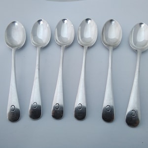 May include: A set of six silver spoons, each with an oval bowl and a long, tapered handle. A small, dark mark is visible near the base of each handle. The spoons are laid out on a pale grey surface.
