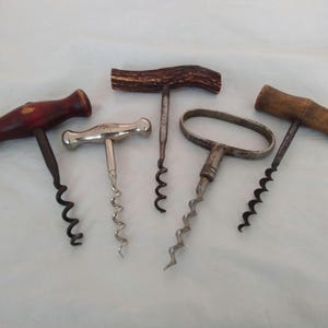May include: Five vintage corkscrews with wooden and metal handles. The corkscrews feature various designs, including a red wooden handle, a silver metal handle, and an antler handle. The corkscrews are displayed on a white surface.