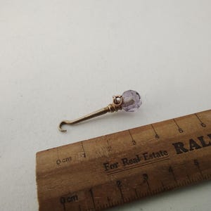 May include: A small, ornate metal tool with a hook and a faceted, light purple crystal bead. The tool is next to a wooden ruler with markings in inches and centimeters.