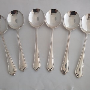 May include: A set of six polished silver-toned spoons arranged in a row. Each spoon features a wide, rounded bowl and a slender handle that widens toward the base, which has a subtle, decorative notched detail at the very end of the handle.