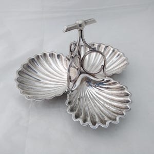 May include: A silver-coloured metal serving dish featuring three interconnected shell-shaped bowls arranged in a clover pattern. A central vertical handle with a horizontal bar at the top connects the three bowls, allowing for easy transport and serving.