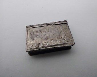 May include: A silver metal matchbox with the words "Blackpool" embossed on the front. The matchbox has a worn, vintage look and features a detailed image of a building.