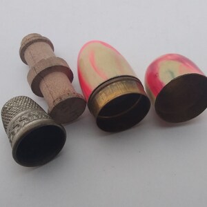 K&ouml;nnte beinhalten: Drei Vintage-N&auml;hzubeh&ouml;rteile: ein silberner Fingerhut, eine Holzspule und zwei kleine, eif&ouml;rmige Beh&auml;lter mit rosa und wei&szlig;en Malereien.