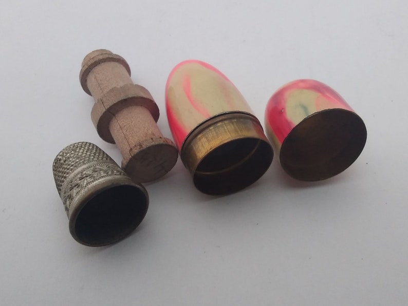 K&ouml;nnte beinhalten: Drei Vintage-N&auml;hzubeh&ouml;rteile: ein silberner Fingerhut, eine Holzspule und zwei kleine, eif&ouml;rmige Beh&auml;lter mit rosa und wei&szlig;en Malereien.
