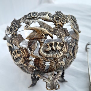 May include: A silver and glass decorative bowl featuring a bird and nest design. The bowl has a delicate, ornate design with floral accents and a clear glass centre.