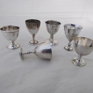 May include: A set of six silver-coloured metal egg cups with a smooth, polished finish. Five of the egg cups are standing upright on circular bases, while one egg cup is lying on its side. Each cup features a rounded bowl and a pedestal stem.