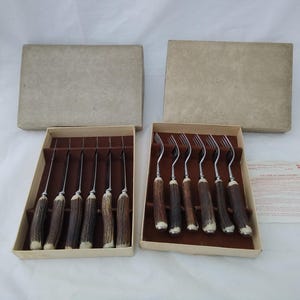 May include: Two rectangular cardboard boxes containing sets of stainless steel cutlery with textured brown antler handles. One box holds six knives, and the other holds six forks. A small white informational card is placed next to the boxes.