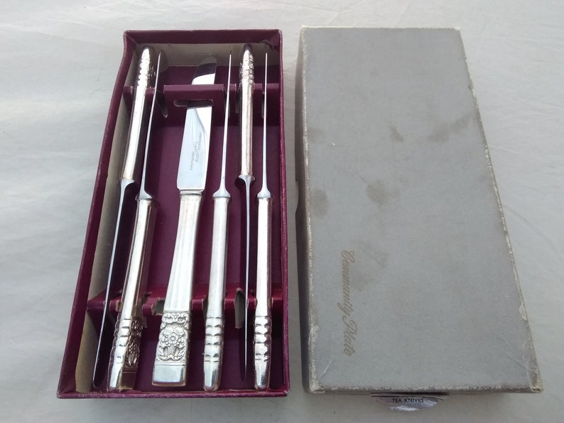Puede incluir: Un juego de cinco cuchillos para carne plateados en una caja color burdeos con las palabras "Community Plate" impresas en la caja. Los cuchillos est&aacute;n en una caja forrada de terciopelo con ranuras individuales para cada cuchillo.