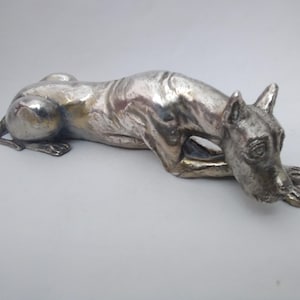 Puede incluir: Una figura de metal en tono plateado de un perro en posición tumbada y de descanso. La escultura presenta una superficie lisa y reflectante con contornos y musculatura visibles a lo largo del lomo y las patas, con la cabeza baja hacia las patas.