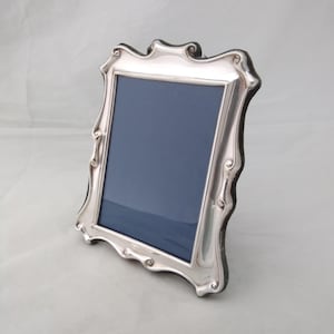 May include: A silver-toned picture frame with a polished, reflective finish and a decorative, undulating border. The frame features a rectangular central opening containing a dark blue insert, set against a neutral, light-colored backdrop.