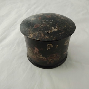 May include: A round black lacquered box with a lid decorated with gold and red floral designs. The box is approximately 7.5 cm in diameter.