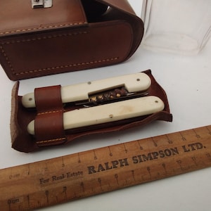 May include: A brown leather case with a silver clasp, holding a wine opener and a knife with cream-coloured handles. A wooden ruler with the text "RALPH SIMPSON LTD." is also visible.