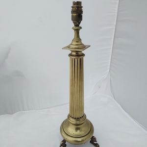 Könnte beinhalten: Eine goldfarbene Tischlampe aus Messing mit einem geriffelten Säulendesign und einem quadratischen Sockel. Die Lampe hat drei Füße, die Tierpfoten ähneln.