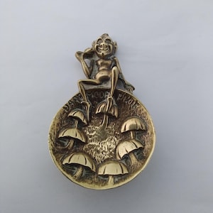 May include: A brass-colored decorative piece featuring a seated pixie figure atop a circular base. The base is adorned with several mushroom-shaped elements. The pixie is smiling and holding an object. The piece has a detailed, textured surface.