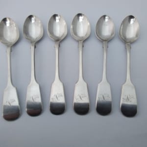May include: A set of six silver-colored metal spoons arranged in a row. Each spoon features a smooth, oval-shaped bowl and a flat, flared handle. The end of each handle is engraved with a decorative monogram consisting of the letters A and V.