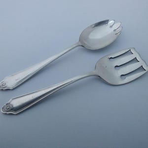 May include: A pair of silver-toned serving utensils consisting of a slotted spoon with a rounded bowl and a wide, flat server with three rectangular cutouts. Both pieces feature a tapered handle with a decorative embossed pattern at the base.