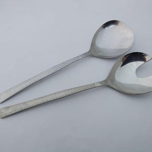 May include: Two silver salad servers. One is a spoon, the other has a slotted design. Both have textured handles. The servers are on a white background. The spoon has a rounded bowl, and the other server has a curved design.