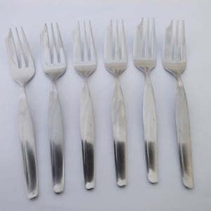 May include: A set of six silver-toned stainless steel forks arranged in a row. Each fork features a four-tined head and a smooth, slightly tapered handle with a polished finish. The forks are identical in design, showing a clean, metallic surface.