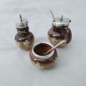 May include: A set of three brown ceramic salt and pepper shakers with silver lids and a spoon. The shakers have a tree design and the lids have a silver finish.