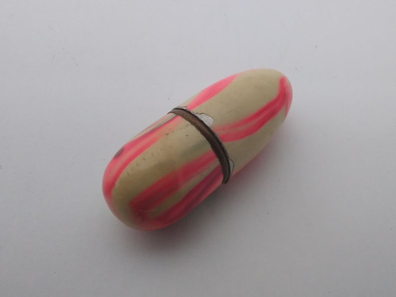 K&ouml;nnte beinhalten: Eine kleine, cremefarbene Pillendose mit rosa Streifen. Die Pillendose ist aus Kunststoff und hat einen Klappdeckel.