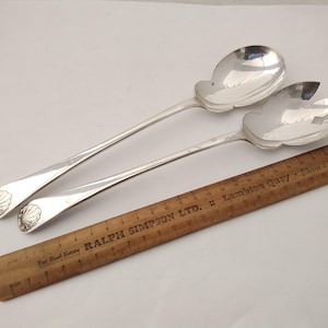 May include: A pair of silver-toned salad serving utensils, including a solid spoon and a three-pronged fork, each featuring a shell motif on the handle, positioned next to a wooden ruler measuring approximately 30 cm in length.