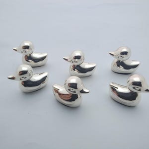 May include: A set of five small, shiny, silver-coloured duck figurines. Each duck is depicted in a smooth, reflective metallic finish with a rounded body, a distinct beak, and a small tail, arranged in a group on a plain, light-coloured surface.