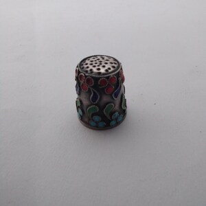 Puede incluir: Un dedal plateado con un diseño floral decorativo en esmalte rojo, azul y verde. El dedal tiene una parte superior perforada y probablemente se utiliza para coser o hacer manualidades. El diseño es simétrico y detallado.