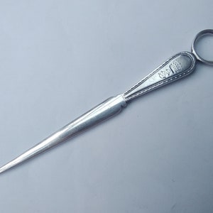 May include: A silver letter opener with a decorative handle and a ring at the top. The handle has an engraved design.