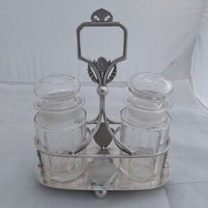 May include: Silver-plated condiment set with two glass jars with lids. The jars have a fluted design and are held in a decorative stand with a handle.