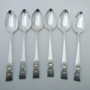 May include: Six silver spoons. Each spoon has an oval bowl, a long, ridged handle, and a decorative floral design at the base. The spoons are reflective, with a bright, shiny appearance. The spoons are arranged in a row.