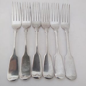 May include: Six silver forks with tines and wide, flat handles. The handles feature a decorative, scalloped design at the base. The forks are arranged in a row against a white background, highlighting their polished, reflective surfaces.