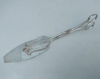 May include: Silver cake server with a decorative handle and a pair of tongs. The server has a long, thin blade and a fork-like design at the end.