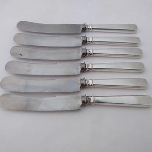 May include: Six silver butter knives arranged in a row. Each knife has a curved blade and a rectangular handle. The knives are made of a shiny, reflective metal, likely stainless steel.