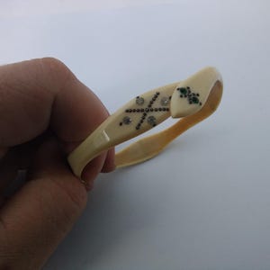 May include: A cream-coloured bangle bracelet shaped like a snake. The head features small green stones for eyes, and the body is decorated with a cross pattern made of tiny dark beads and four clear, sparkling rhinestones set into the smooth surface.