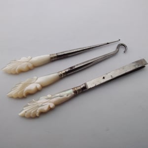May include: Three vintage grooming tools featuring carved cream-coloured handles with a leaf-like pattern. The set includes a pointed metal pick, a curved button hook, and a pair of flat metal tweezers, each with a metallic ferrule connecting the handle.