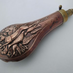 Könnte beinhalten: Eine Pulverflasche aus Kupfer mit einem geprägten Design einer Jagdszene mit einem Hund und Vögeln. Die Flasche hat einen Messingverschluss und eine Schlaufe zum Tragen.