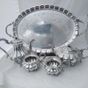 May include: A silver tea set comprising an oval tray, two teapots, a creamer, and a sugar bowl. The set features intricate detailing and handles. The tray has scalloped edges and handles. The tea set is displayed on a white surface.