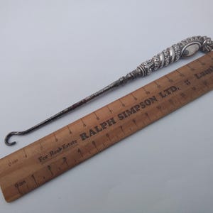 May include: Silver shoe horn with an ornate handle. The handle is decorated with a swirling pattern and a small, round, silver plate. The shoe horn is 30.5 cm long.