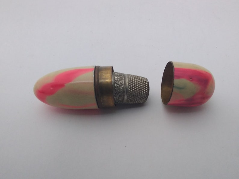 K&ouml;nnte beinhalten: Ein wei&szlig;es und rosafarbenes N&auml;hk&auml;stchen mit einem silbernen Fingerhut darin. Das K&auml;stchen ist ge&ouml;ffnet und der Fingerhut ist sichtbar.