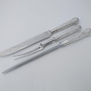 May include: A silver carving set featuring a long knife, a two-pronged fork, and a sharpening steel. The handles have a decorative floral design. The set is displayed on a white surface.