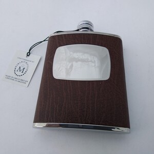 May include: A brown leather-effect flask with a silver trim and cap. The flask features a rectangular, recessed panel on the front. A tag with the text "Craftsmanship by Marlborough" is attached.