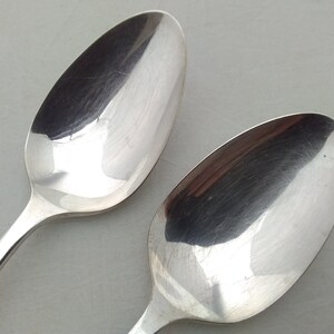 May include: Two silver spoons with a shiny, polished finish. The spoons are laying on a white surface.