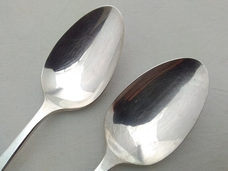 May include: Two silver spoons with a shiny, polished finish. The spoons are laying on a white surface.
