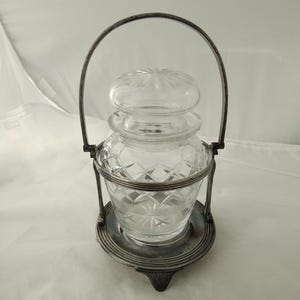 May include: A clear glass jar with a lid, set in a silver-colored metal frame with a handle. The jar has a diamond pattern and sits on a matching base. The handle arches over the jar.