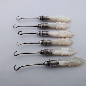 May include: A set of six button hooks arranged horizontally. Each tool features a curved metal hook, a decorative silver-toned metal collar with floral engravings, and a carved, off-white, pearlescent handle with a leaf-like pattern.